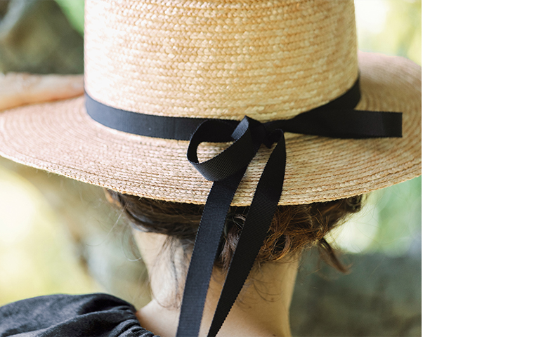 田中帽子店 滝口和代さん×田中帽子店コラボ quiet straw hat の後ろ姿ディテール。黒いリボンを後ろで結んだ麦わら帽子をかぶった人物の後ろ姿。自然光に照らされ、編み目の美しい質感とリボンの柔らかな動きが際立つカット。