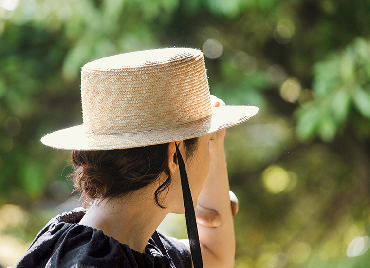田中帽子店 滝口和代さん×田中帽子店コラボ quiet straw hat の着用ディテール。黒いリボン付きの麦わら帽子をかぶった人物が、緑の木々の中で帽子のつばに手を添える横向きカット。自然光に照らされた編み目の質感が美しく、穏やかな雰囲気を感じるイメージ。