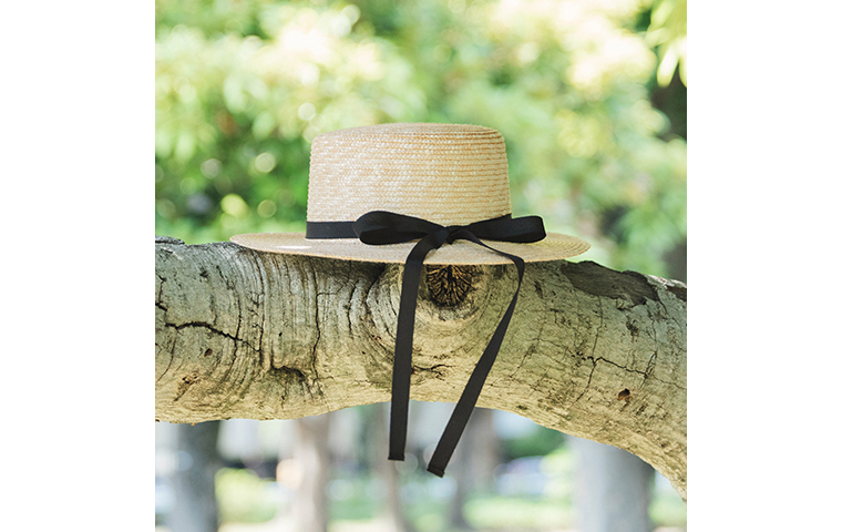 田中帽子店 滝口和代さん×田中帽子店コラボ quiet straw hat の商品ディテール。黒いリボンを結んだ麦わら帽子が木の枝に掛けられている様子。自然光に照らされた編み目の美しい質感と、ナチュラルな雰囲気が際立つイメージカット。