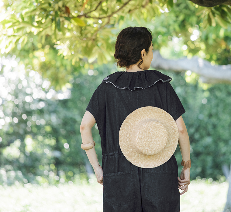 田中帽子店 滝口和代さん×田中帽子店コラボ quiet straw hat の着用イメージ。黒いリボン付きの麦わら帽子を背中に掛けた人物の後ろ姿。黒のワンピースと自然光に包まれた緑の木々が調和し、穏やかな夏の雰囲気を感じるスタイリングカット。