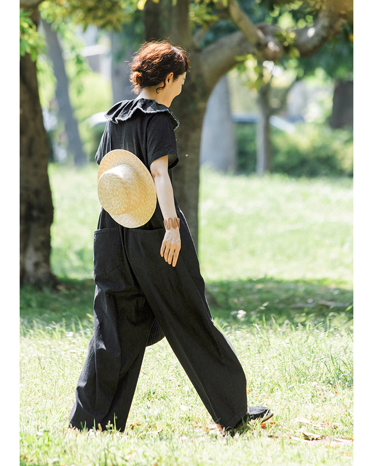 田中帽子店 滝口和代さん×田中帽子店コラボ quiet straw hat の着用イメージ。黒いワンピースを着た人物が、麦わら帽子を背中に掛けて緑の木々の中を歩く後ろ姿。自然光に包まれた穏やかな雰囲気と、軽やかな動きが感じられるスタイリングカット。
