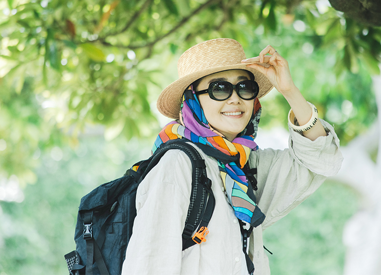 田中帽子店 滝口和代さん×田中帽子店コラボ quiet straw hat の着用イメージ。麦わら帽子にカラフルなスカーフを合わせ、サングラスとリネンシャツを着た人物が屋外で微笑む姿。黒いバックパックを背負い、自然光に包まれた緑の木々の下で軽やかな雰囲気を感じるスタイリングカット。
