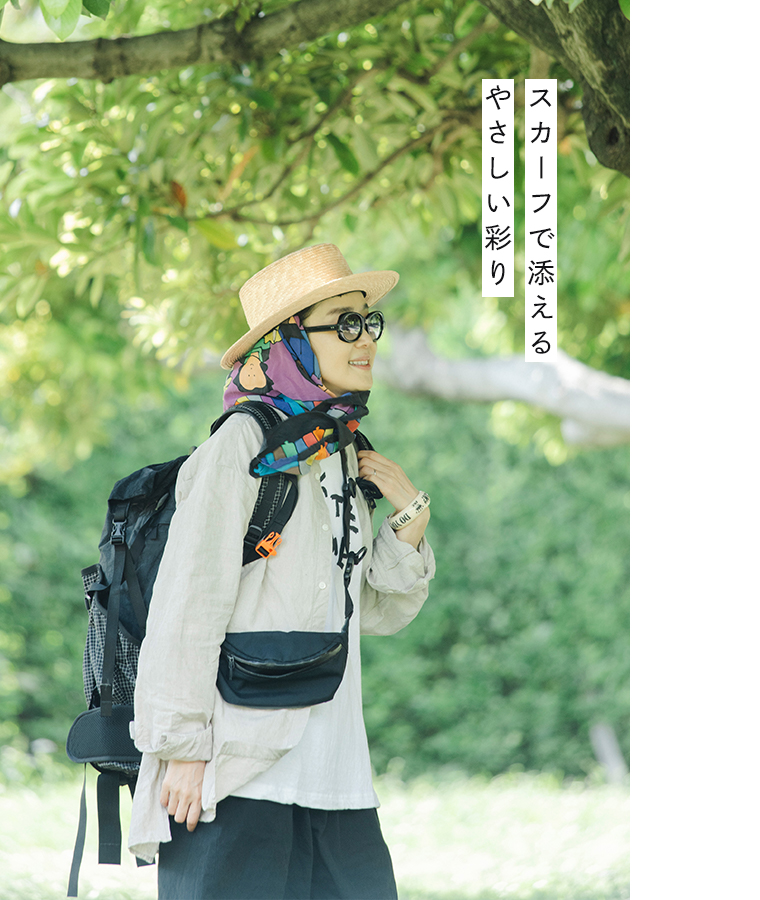 田中帽子店 滝口和代さん×田中帽子店コラボ quiet straw hat の着用イメージ。麦わら帽子にカラフルなスカーフを合わせ、サングラスとリネンシャツを着た人物が屋外で微笑む姿。黒いバックパックを背負い、緑の木々の下で軽やかに歩くスタイル。「スカーフで添える やさしい彩り」の文字入りで、ナチュラルなフェススタイルを表現したカット。