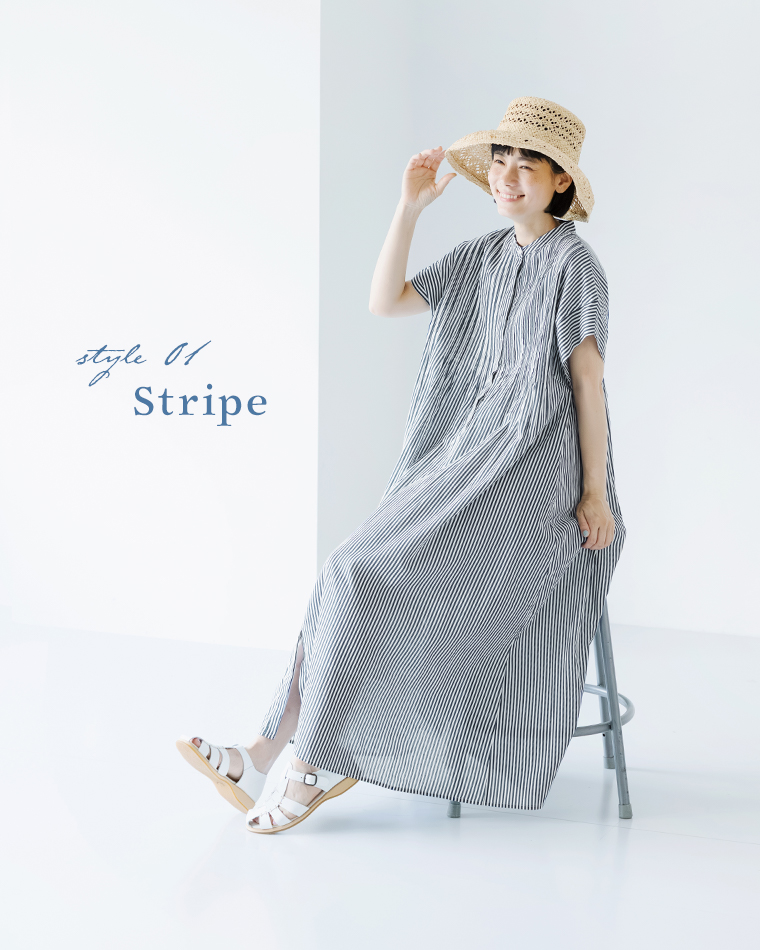 style 01 Stripeのコーディネート。ネイビーストライプの前開きピンタックワンピースを着て、麦わら帽子と白のサンダルを合わせた女性が、丸椅子に腰掛けて笑顔で帽子に手を添えている全身カット。