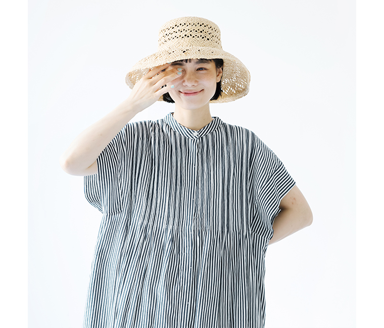 Noiaキャンブリックボイル前開きピンタックワンピース（ネイビーストライプ）と麦わら帽子を着用した女性が、右目でウィンクをしながら、左手を目のあたりに添えている上半身のカット。