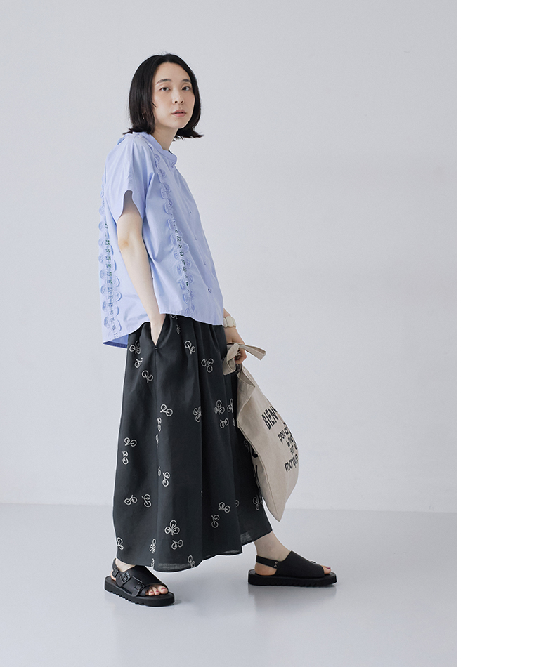 nop de nod コットンスタンドカラーバテンレースシャツ（ブルー）に、刺繍入りの黒いボリュームスカートとサンダルを合わせたモデルの斜め横姿・全身コーディネート画像。左手にトートバッグを持っています。