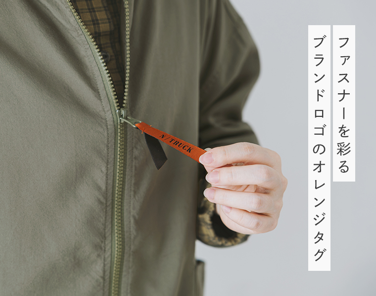 カーキのスタンドカラージャケットのファスナーに付いた、オレンジのブランドロゴタグを見せたディテール画像