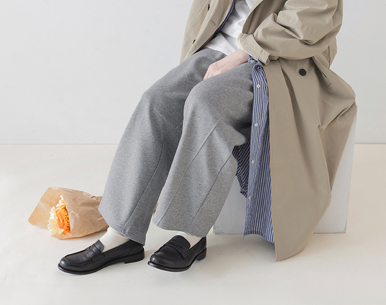 杢グレーのこなれスウェットカーブパンツにベージュのロングコートと黒のローファーを合わせた座り姿の足元アップ