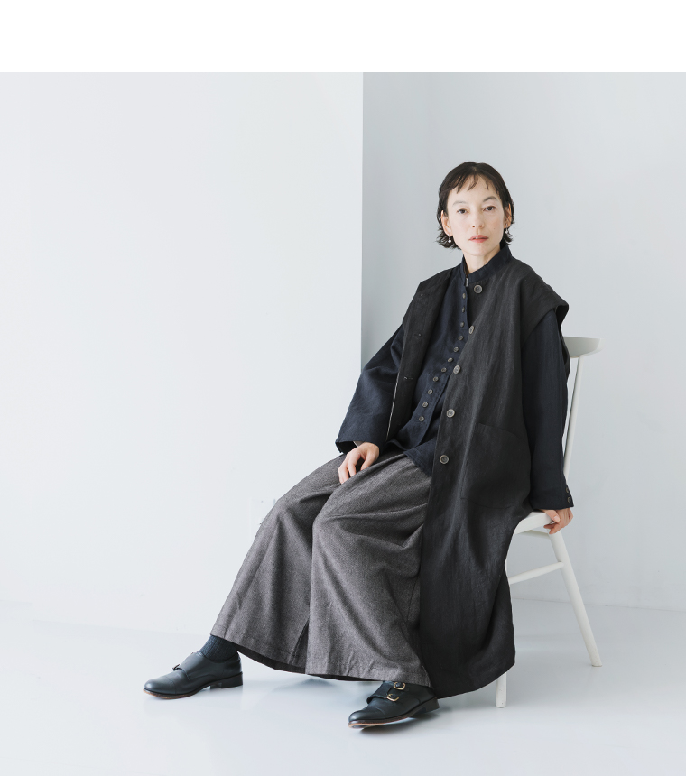 ブラックのジャケットとロングベストを着用して椅子に腰掛ける女性モデル