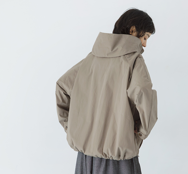 グレーベージュのフードZIPジャケットを着用し、丸みのあるシルエットと裾デザインを見せたバックスタイル
