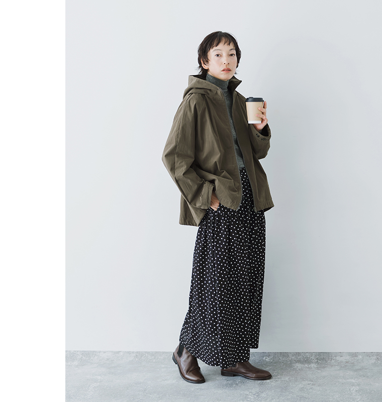 カーキグリーンのフードZIPジャケットをドット柄ロングスカートに合わせ、カップを持ったモデルの全身コーディネート