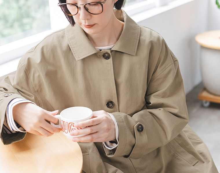 ベージュのわたしの定番ステンカラーコートを着用し、カップを手に持つ上半身カット