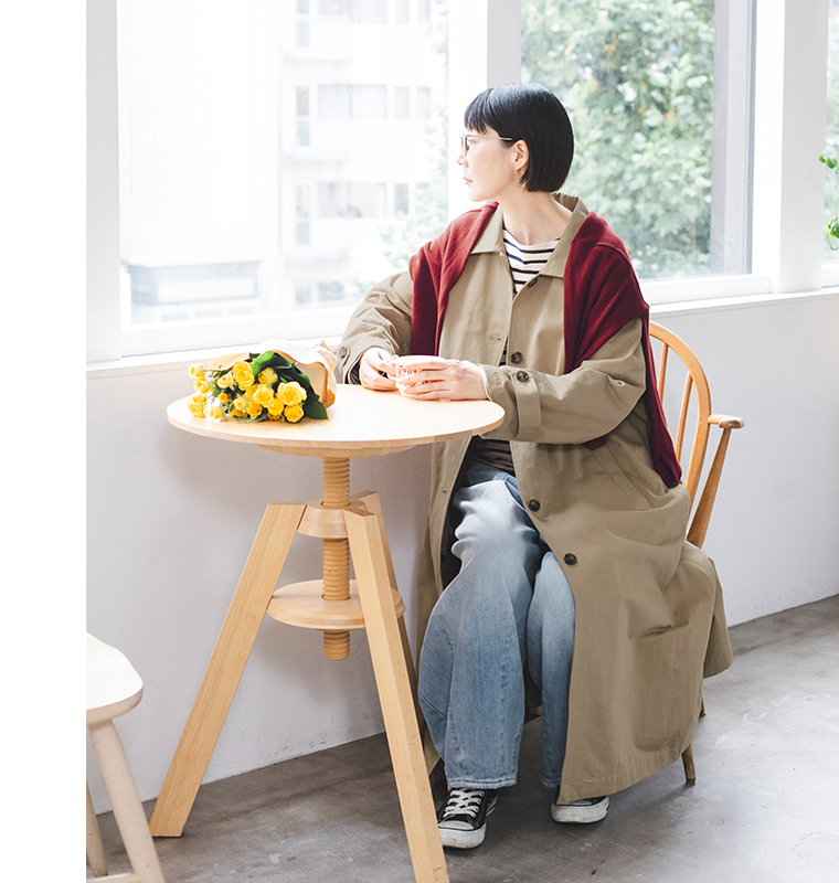 ベージュのわたしの定番ステンカラーコートを着用し、赤いニットを肩掛けした座り姿のコーディネート
