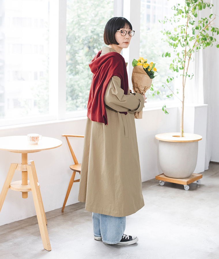 ベージュのわたしの定番ステンカラーコートに赤いニットを肩掛けし、花束を持った横向きの着用カット
