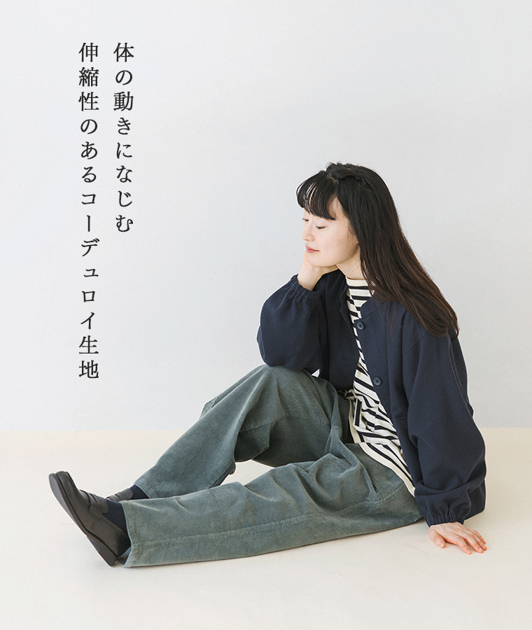 コーデュロイワークストレートパンツのグリーンをネイビーのジャケットとボーダートップスに合わせたモデルの座り姿