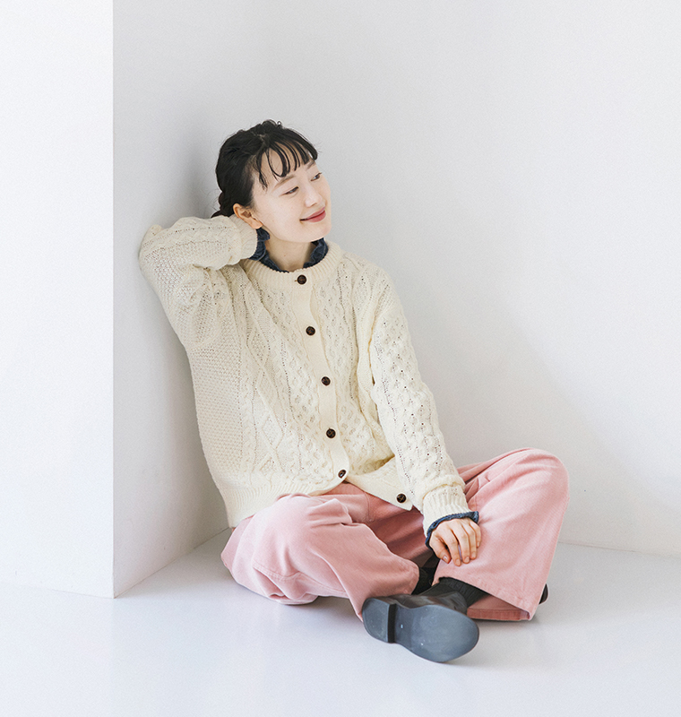 ケーブルニットカーディガンをデニムシャツとピンクのパンツに合わせ、床に座るモデルの全身コーディネート