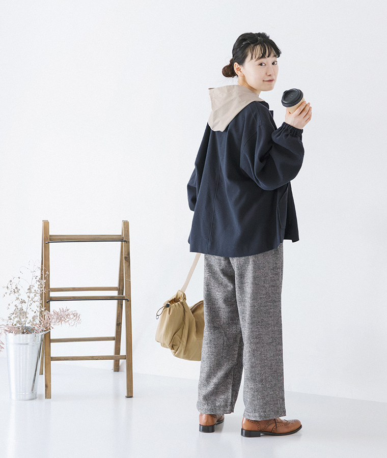 タックツイードパンツにネイビーのジャケットとベージュのフード付きトップスを合わせ、コーヒーカップを手に振り返るモデルの全身コーディネート
