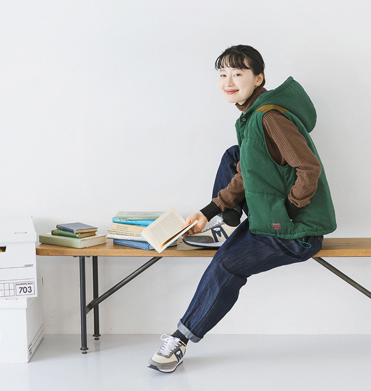 フード付き中わたベストにボーダートップスとデニムパンツを合わせ、ベンチに座って本を読むモデルの全身コーディネート